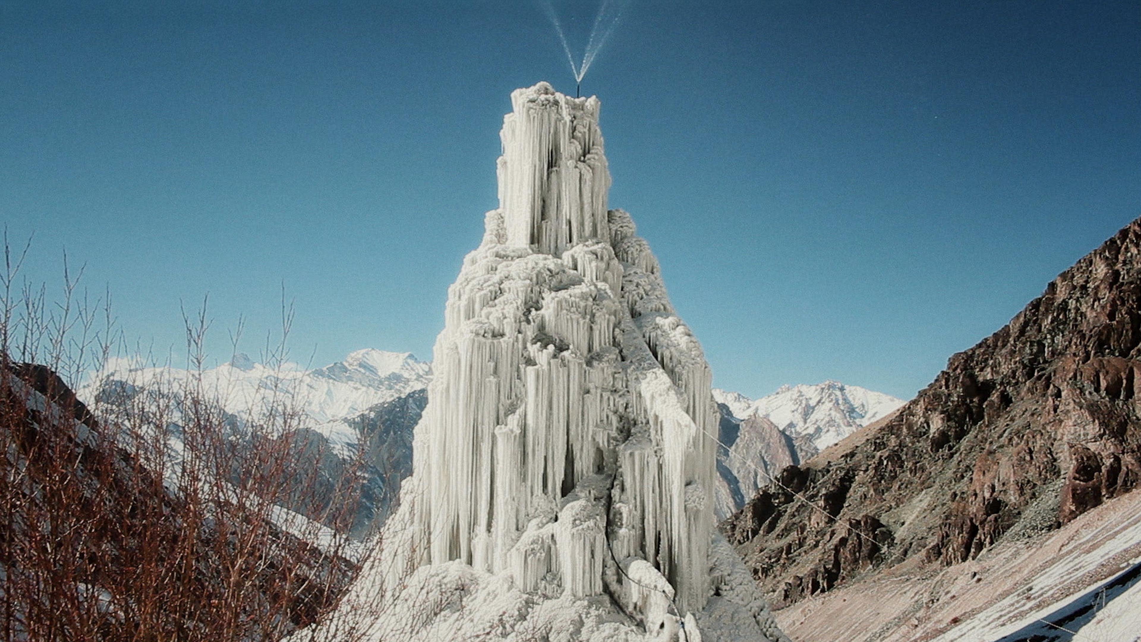A large, artificial ice structure in a mountainous landscape with clear blue sky in the background.