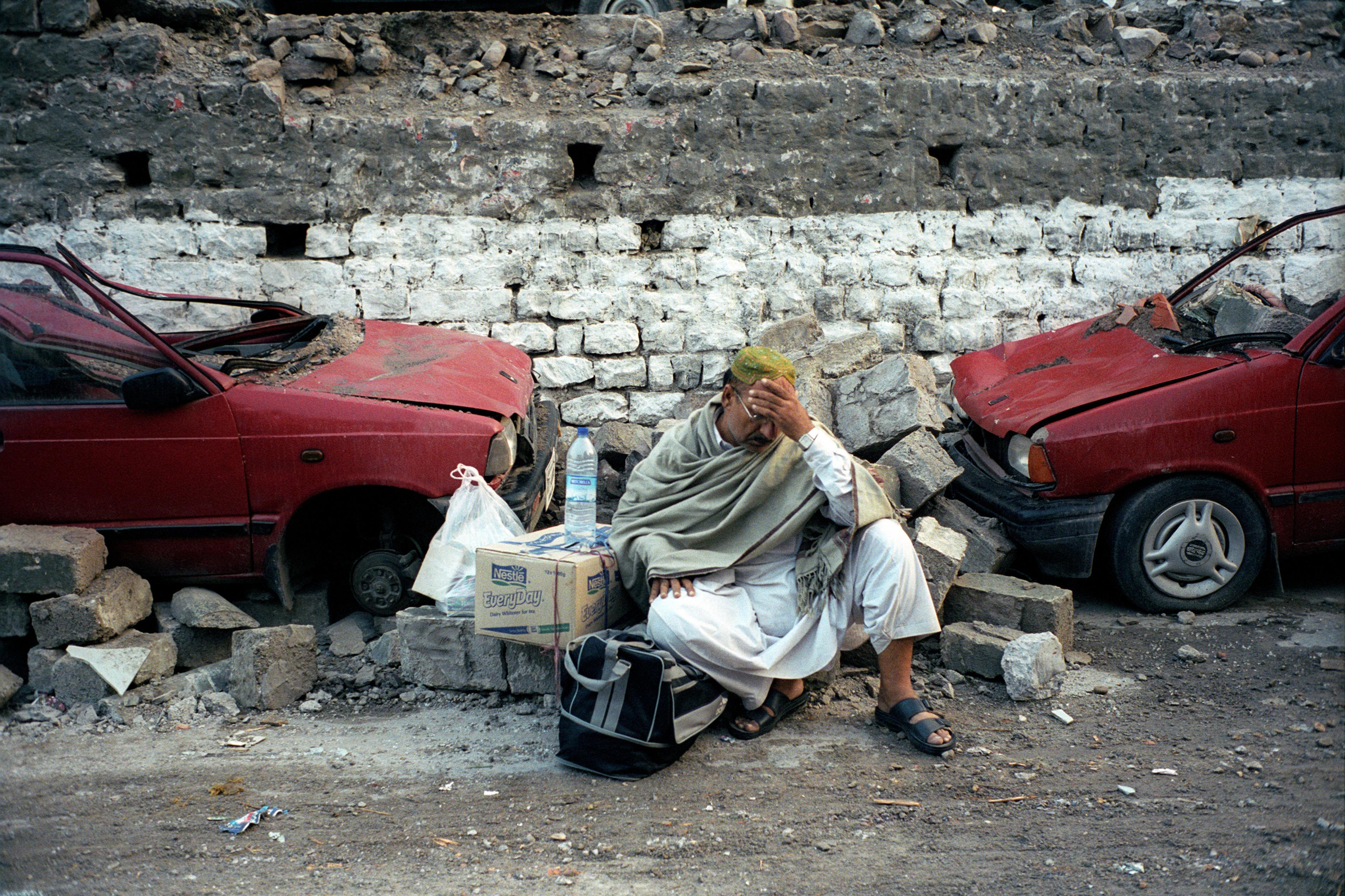 A man wearing a robe sitting amidst rubble between two damaged red cars. He holds his head and has boxes and bags beside him.