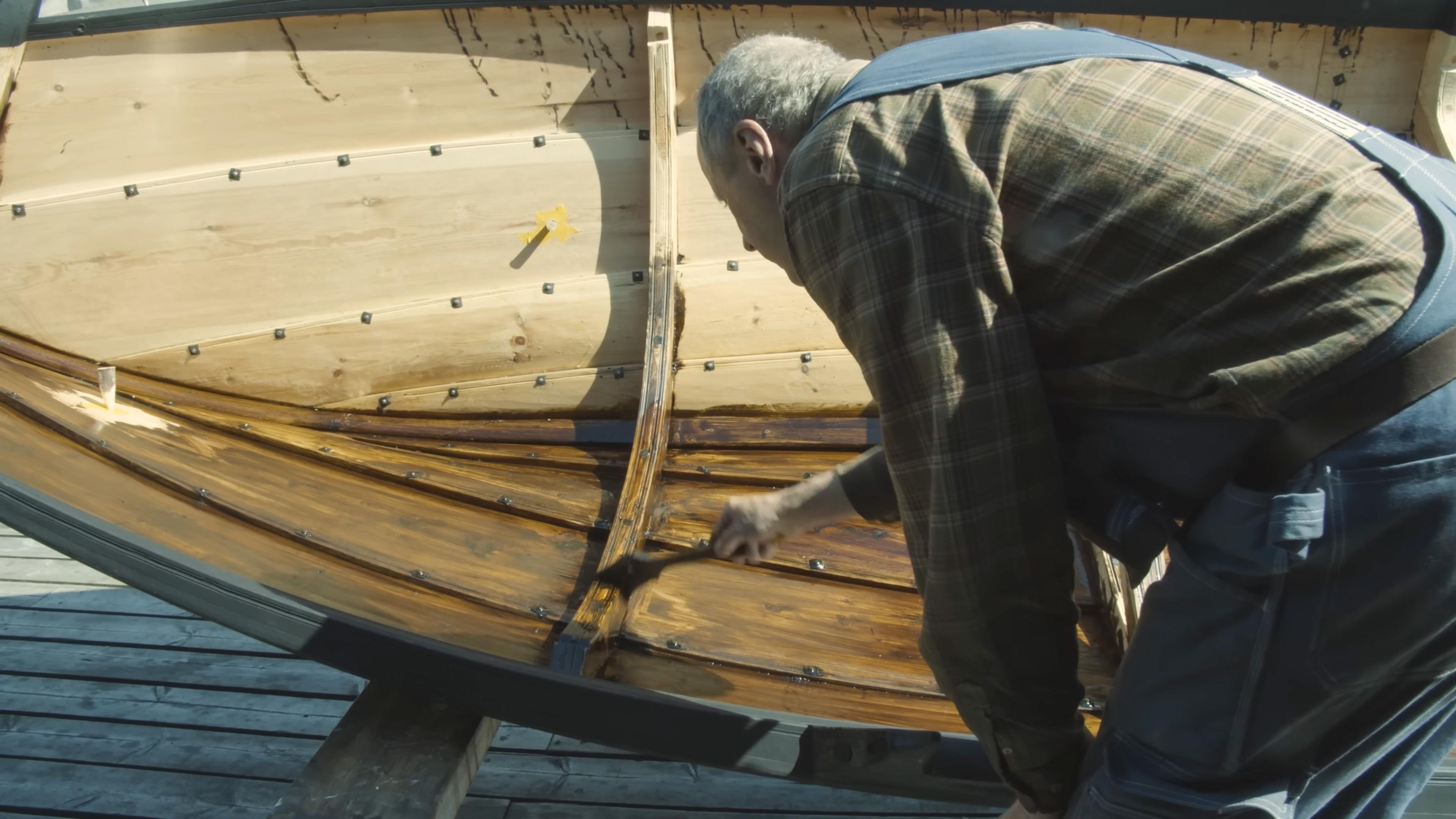 Photo of a person applying varnish to the interior planks of a wooden boat on a sunny day.
